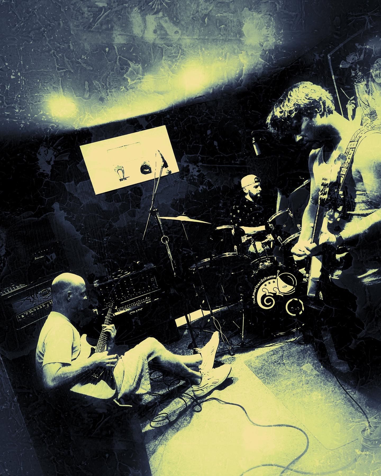 The American Scheme playing live in the practice room: guitarist seated on the floor, drummer behind the kick drum with the band logo, guitarist standing at right