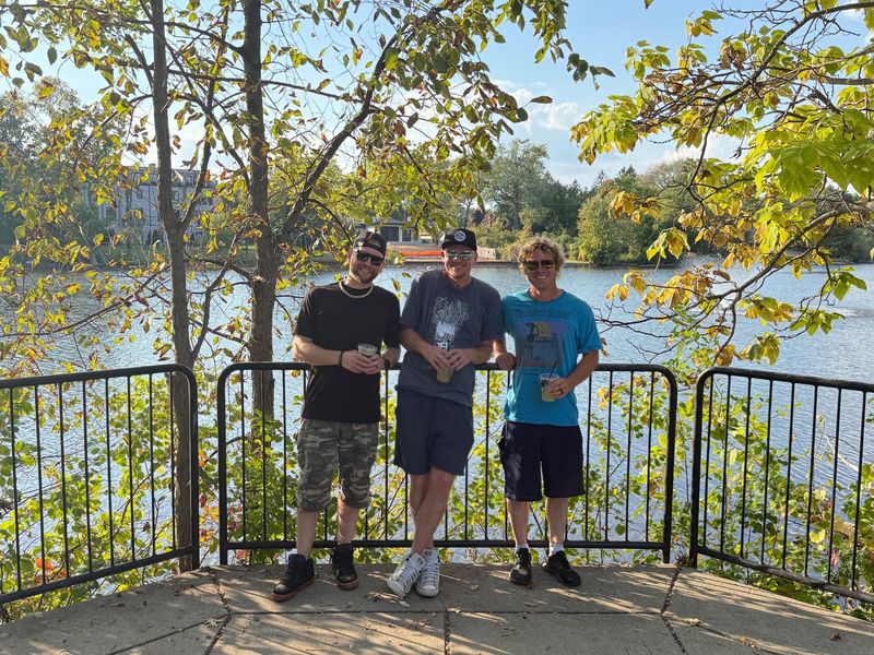 The three band members standing at a waterside railing, trees behind them